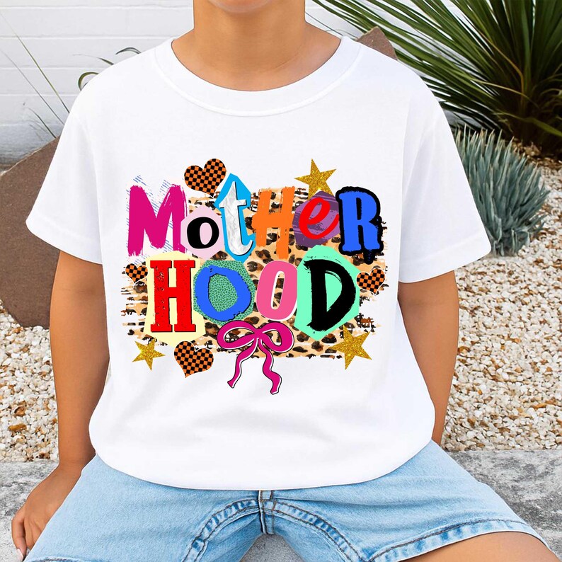 May include: White t-shirt with the word "MOTHERHOOD" in colorful, playful letters. The design includes hearts, stars, and a bow, all set against a leopard print background. A fun and vibrant graphic tee.