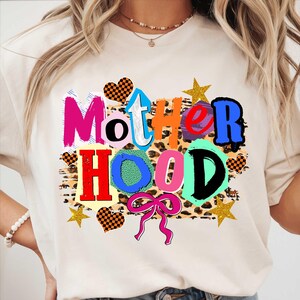 May include: Cream-colored t-shirt with the word "Motherhood" in a colorful, playful design. The word is composed of various colors and fonts, with a leopard print background and decorative hearts, stars, and a pink bow.