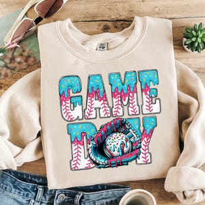 以下が含まれることがあります： ベージュ色のスウェットシャツ。青、ピンク、白のデザインで「GAME DAY」の文字が描かれています。デザインには野球グローブとスプリンクル付きの野球ボールが含まれています。柔らかい素材で作られたスウェットシャツです。