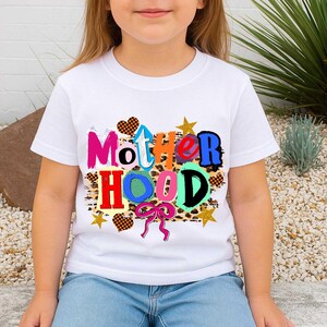 May include: White t-shirt with the word "Motherhood" in colorful, playful lettering. The design includes hearts, stars, and a bow, with a leopard print background. The shirt is worn by a child.