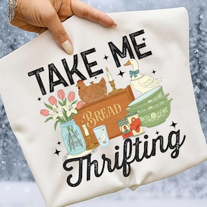 Puede incluir: Camiseta gr&aacute;fica color crema con las palabras negras "TAKE ME Thrifting". El dise&ntilde;o incluye ilustraciones de una gallina, un pato, flores, cuencos, velas y conservas. La camiseta est&aacute; sujeta por una mano con esmalte de u&ntilde;as dorado brillante.