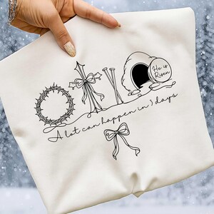 May include: Off-white tote bag featuring a black line drawing of a crown of thorns, a cross with a bow, nails, and an empty tomb with the words "He is Risen." The text "A lot can happen in 3 days" is also included.