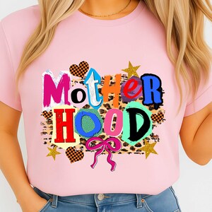 May include: A light pink t-shirt with the word "MOTHERHOOD" in a colorful, patchwork design. The letters are in various colors and patterns, including leopard print, hearts, and stars. A pink bow is at the bottom.