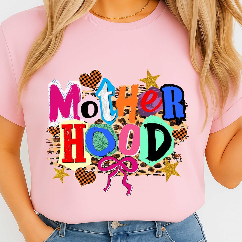 May include: A light pink t-shirt with the word "MOTHERHOOD" in a colorful, patchwork design. The letters are in various colors and patterns, including leopard print, hearts, and stars. A pink bow is at the bottom.