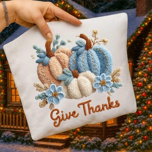 May include: A white decorative pillow featuring embroidered pumpkins in peach, blue, and white, with blue floral accents. The phrase "Give Thanks" is embroidered in brown script, perfect for fall decor.