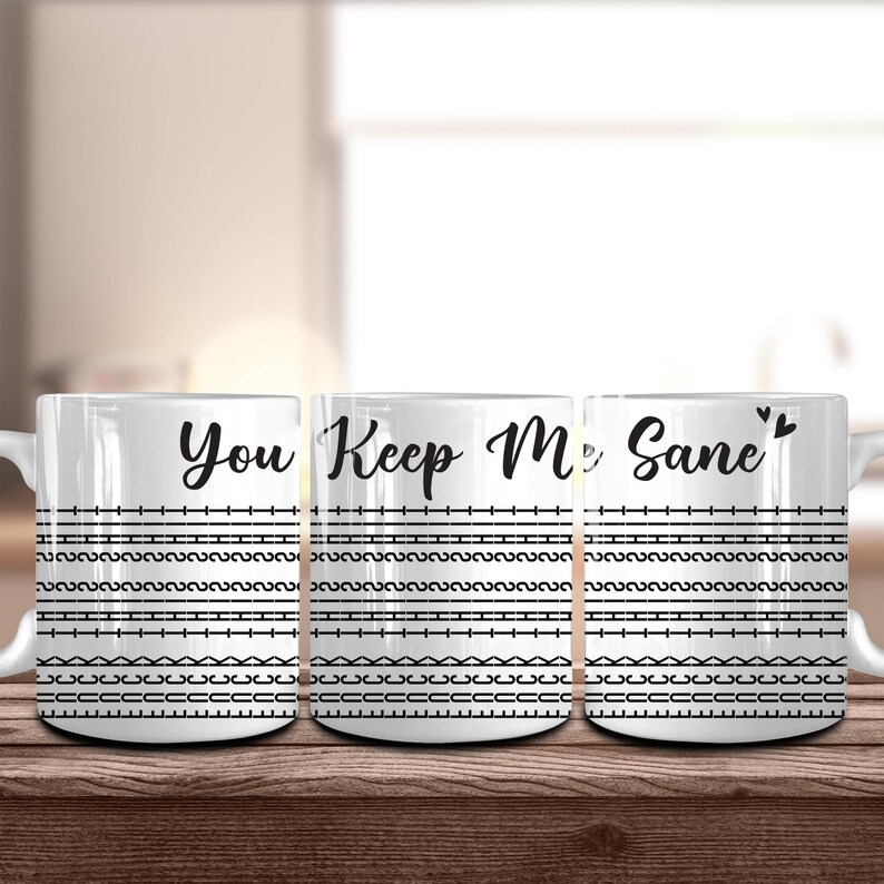 May include: White ceramic mugs with the phrase "You Keep Me Sane" in black script. The mugs feature black horizontal lines and geometric patterns. The mugs are on a wooden surface.