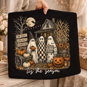 Puede incluir: Una camiseta negra con un gráfico de tres fantasmas frente a una casa con luna llena, calabazas y tallos de maíz. El texto dice Hay Rides, Corn Maze, Pumpkins, Boo Y'all y Tis the season en una fuente de estilo manuscrito.