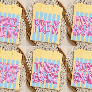 May include: Six yellow t-shirts featuring a light blue vertical striped background and pink text. The shirts display the words Kindergarten, Pre-K, First Grade, Second Grade, Third Grade, and Fourth Grade in a playful, rounded font style.