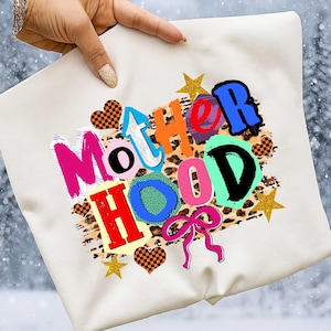 May include: Cream-colored sweatshirt with the word "MOTHERHOOD" in colorful, layered text. The design includes leopard print, stars, hearts, and a pink bow. The sweatshirt is held up by a hand.