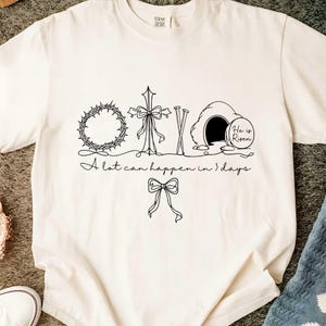 May include: Cream-colored t-shirt with a black line drawing of a crown of thorns, cross, nails, and an empty tomb with the words "He is Risen." The text "A lot can happen in 3 days" is also included.