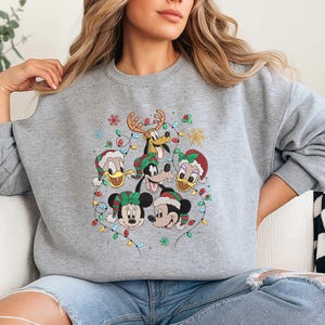 Könnte beinhalten: Ein graues Sweatshirt mit einem festlichen Design mit Disney-Figuren, darunter Mickey und Minnie Mouse, Donald und Daisy Duck sowie Goofy, die alle Weihnachtsmützen tragen und von bunten Lichtern umgeben sind.