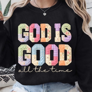 May include: Black sweatshirt with the words "GOD IS GOOD" in colorful patchwork letters. The phrase "all the time" is written in a cursive font below the main text. The letters have floral and gingham patterns.