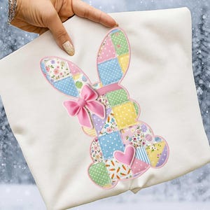 May include: A white cushion featuring a patchwork bunny design. The bunny has a pink bow, a pink heart, and various pastel-colored patterned squares. The design is outlined in pink.