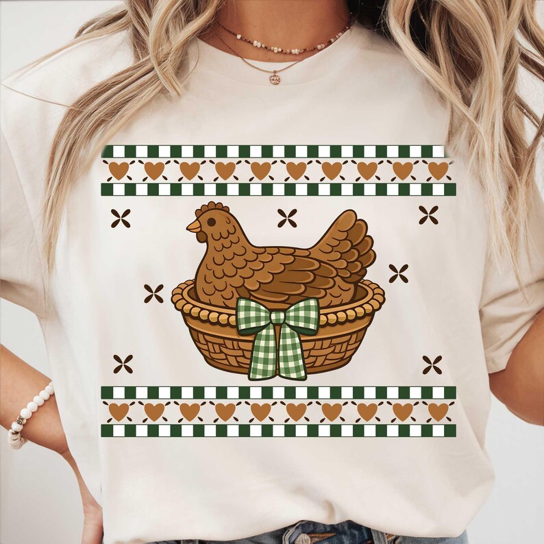 May include: Cream-coloured t-shirt featuring a brown hen in a woven basket with a green and white checkered bow. The design includes brown hearts, green and white checkered patterns, and brown cross shapes.