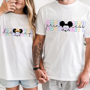 以下が含まれることがあります： 虹色のグラフィックデザインが施された白いTシャツを着たカップル。女性のTシャツには「ディズニー プリンセス」と、王冠とサングラスをかけたミニーマウスの漫画の画像が描かれています。男性のTシャツには「セキュリティ」と、サングラスをかけたミッキーマウスの漫画の画像が描かれています。
