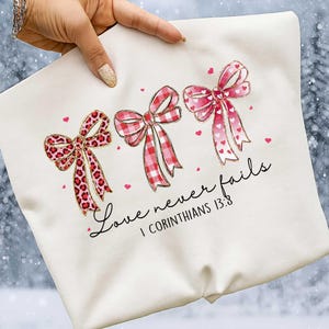 Op de afbeelding: Crèmekleurige sweatshirt met drie decoratieve strikken in rode en roze patronen, waaronder luipaardprint, ruitjes en harten. De tekst "Love never fails" en "I Corinthians 13:8" staat eronder.