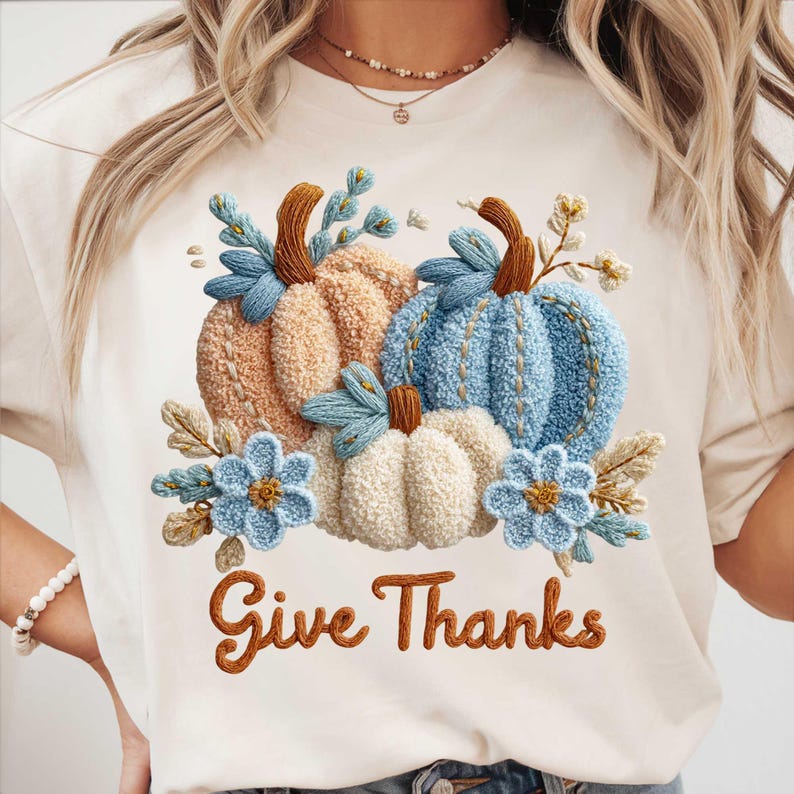 May include: Cream-colored t-shirt featuring an embroidered design of pumpkins and flowers in shades of orange, blue, and white. The phrase "Give Thanks" is written in brown script below the design. The shirt has a crew neck and short sleeves.