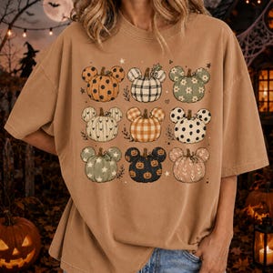 May include: A tan t-shirt featuring a grid of nine pumpkin illustrations, each shaped like mouse ears. The pumpkins display various patterns including polka dots, plaid, gingham, stars, and small jack-o'-lantern faces, accented by tiny stars.