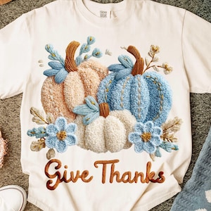 May include: Cream-colored t-shirt featuring an embroidered design of three pumpkins in shades of peach, blue, and white, with blue floral accents. The phrase "Give Thanks" is embroidered in a brown script across the bottom. A cozy fall-themed apparel item.
