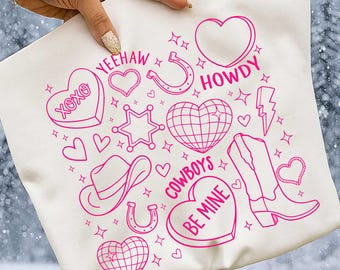 May include: Off-white tote bag with a pink cowboy-themed design. The design includes hearts, a cowboy hat, horseshoe, boots, and the phrases "Yeehaw," "Howdy," and "Cowboys Be Mine." The bag is held by a person.