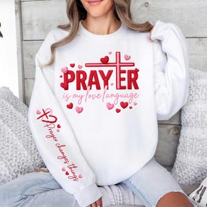 Peut inclure: Sweat-shirt blanc avec l'inscription "PRAYER is my love language" en rouge et un motif de croix rouge. La manche porte l'inscription "Prayer changes things" en rouge. Des cœurs roses et rouges sont dispersés autour du motif.