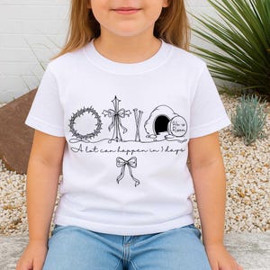 May include: White t-shirt with a black line drawing of a crown of thorns, cross, tomb, and bow. The text "A lot can happen in 3 days" is below the drawings. The words "He is Risen" are inside the tomb.