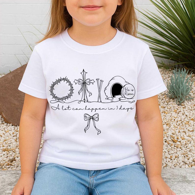 May include: White t-shirt with a black line drawing of a crown of thorns, cross, tomb, and bow. The text "A lot can happen in 3 days" is below the drawings. The words "He is Risen" are inside the tomb.