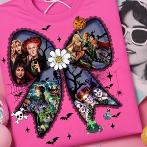 Op de afbeelding: Een roze t-shirt met een groot strikdesign met afbeeldingen van heksen en Halloween-thema's. De strik is versierd met vleermuizen, sterren en een madeliefje. Een pompoen en een schedel zijn ook zichtbaar.