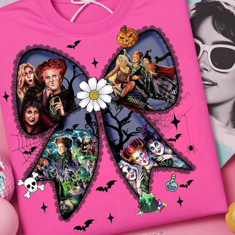 Op de afbeelding: Een roze t-shirt met een groot strikdesign met afbeeldingen van heksen en Halloween-thema's. De strik is versierd met vleermuizen, sterren en een madeliefje. Een pompoen en een schedel zijn ook zichtbaar.
