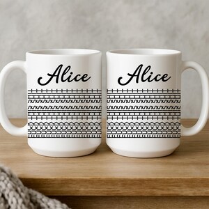 May include: Two white ceramic mugs with the name "Alice" in black script. Each mug features a black geometric pattern around the middle. The mugs are on a wooden surface.