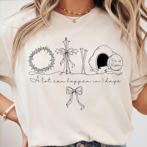 May include: Cream-colored t-shirt featuring a black line drawing of a crown of thorns, cross, nails, and an empty tomb with the words "He is Risen." The text "A lot can happen in 3 days" is written below the illustrations.