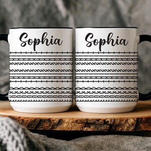 May include: Two white ceramic mugs with black handles and rims. Each mug features the name "Sophia" in a cursive font, above a black and white geometric pattern. The mugs are displayed on a wooden surface.