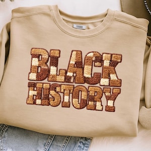 May include: A tan sweatshirt with the words "BLACK HISTORY" in a brown, checkered, and textured design. The sweatshirt has a crew neck and long sleeves. The overall style is casual and comfortable.