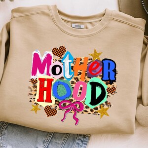 May include: A tan sweatshirt with the word "Motherhood" in colorful, block letters. The design includes hearts, stars, and a leopard print background. A pink bow is at the bottom of the design.