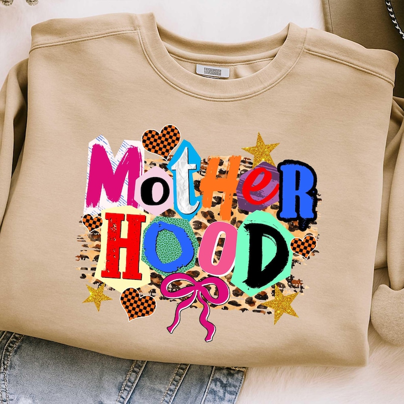 May include: A tan sweatshirt with the word "Motherhood" in colorful, block letters. The design includes hearts, stars, and a leopard print background. A pink bow is at the bottom of the design.