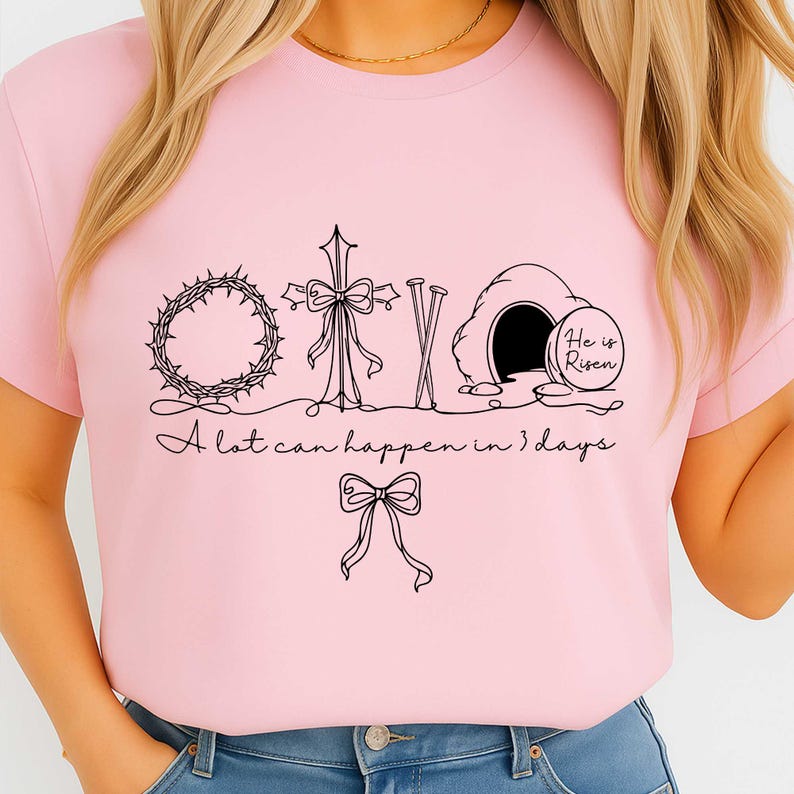 May include: A light pink t-shirt featuring a black graphic design with religious symbols, including a crown of thorns, a cross, and an empty tomb. The text "A lot can happen in 3 days" is also included.