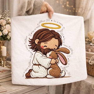 以下が含まれることがあります： イエスが茶色のウサギを抱きしめている漫画の刺繍が施された白い布。イエスの頭上には金の光輪があります。「マタイ28:6 彼は復活した」というテキストが画像を囲んでいます。