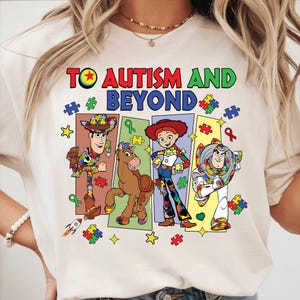May include: Cream-colored t-shirt with the text "TO AUTISM AND BEYOND" in red, green, and blue. The shirt features cartoon characters with puzzle piece designs, including a cowboy, a horse, a cowgirl, and an astronaut.