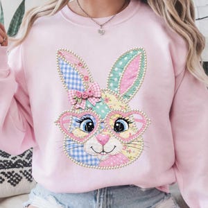 Op de afbeelding: Zachtroze sweatshirt met een patchwork konijn ontwerp. Het konijn heeft hartvormige bril, blauwe ogen en een lachende mond. De oren zijn versierd met ruitjes, stippen en bloempatronen. Een roze strik met rozen siert het hoofd van het konijn.