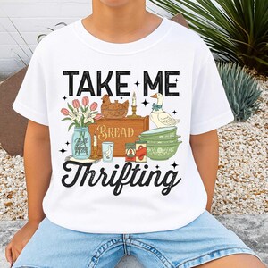 Puede incluir: Camiseta blanca con la frase "TAKE ME Thrifting" en negro. El dise&ntilde;o incluye un tarro de cristal con flores, una caja de pan de madera, una gallina, un ganso, velas y cuencos verdes.