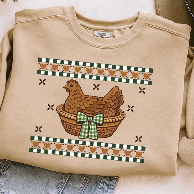 May include: Beige sweatshirt with a brown hen in a woven basket, adorned with a green and white checkered bow. The design includes green and white checkered stripes, brown hearts, and black X's. A charming and cosy design.