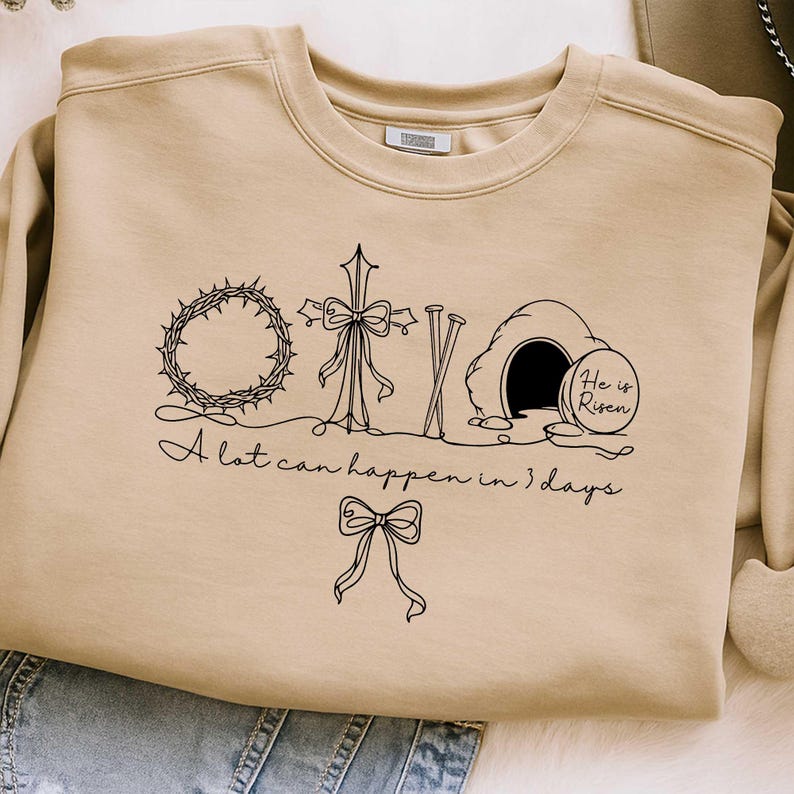 May include: Beige sweatshirt with a black graphic featuring a crown of thorns, cross, nails, and a tomb with the text "He is Risen." The phrase "A lot can happen in 3 days" is written below the graphic.