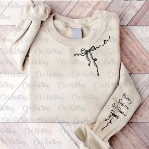 May include: A beige sweatshirt with the word "mama" written in cursive with a bow above it. The sweatshirt has the names "Harry", "Susan", and "William" written on the sleeve with a bow below the names.