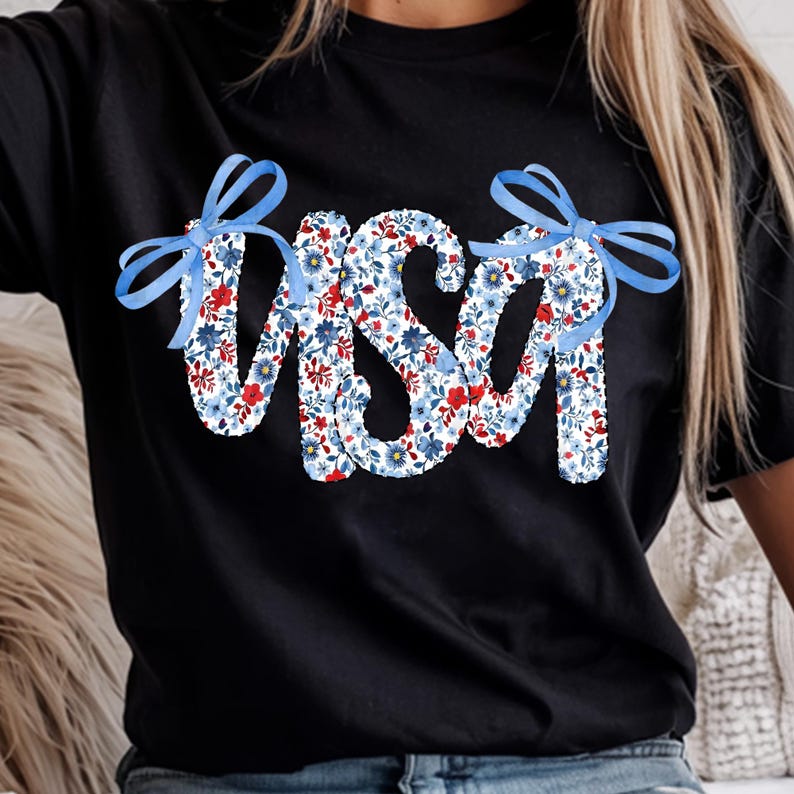 May include: Black t-shirt with the word "USA" in a floral design of red and blue flowers on a white base. The letters are decorated with blue ribbon bows, creating a patriotic theme.