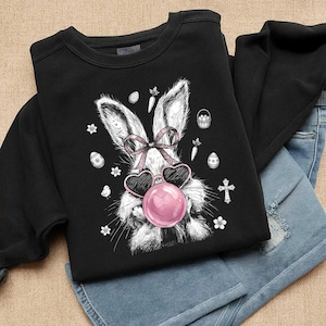 Op de afbeelding: Zwart sweatshirt met een wit en grijs konijn met een hartvormige zonnebril en een roze bel. Het ontwerp bevat paas-elementen. Het sweatshirt wordt gecombineerd met een blauwe jeans.