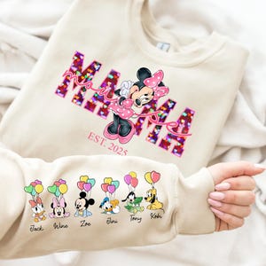 Puede incluir: Una sudadera beige claro con un gráfico rosa "MAMA" con Minnie Mouse y corazones. La sudadera tiene el texto "EST. 2025" debajo del gráfico. La manga tiene una fila de seis personajes de dibujos animados con globos y nombres: Jack, Wine, Zoe, Jini, Tony, Kobi.