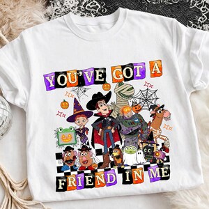 May include: A white t-shirt featuring a graphic print of various animated characters dressed in Halloween costumes. The text "YOU'VE GOT A FRIEND IN ME" is printed in colorful, blocky letters above and below the characters, surrounded by spiderwebs and pumpkins.