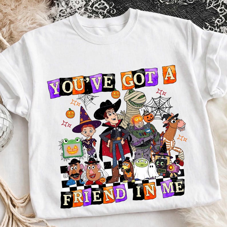 May include: A white t-shirt featuring a graphic print of various animated characters dressed in Halloween costumes. The text "YOU'VE GOT A FRIEND IN ME" is printed in colorful, blocky letters above and below the characters, surrounded by spiderwebs and pumpkins.