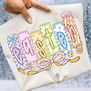 以下が含まれることがあります： クリーム色のイースターをテーマにしたスウェットシャツ。カラフルで装飾的な文字で「EASTER」と書かれています。各文字は異なる色とパターンで、リボンとイースターエッグのグラフィックが描かれています。ニンジンとバスケットも表示されています。