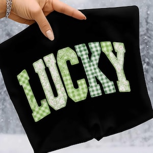 May include: Black fabric with the word "LUCKY" in large, green letters. The letters have a patchwork design, with some sections featuring a gingham pattern and others a floral print. The fabric is held up by a hand.